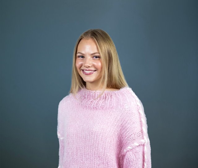 Jente med lyst hår i hestehale og rosa strikket genser smiler bredt mens hun ser rett i kamera