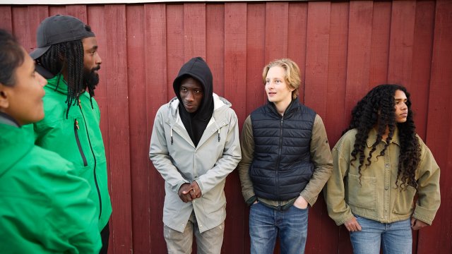 To ungdomsarbeidere i grønne jakker står ved en vegg sammen med tre ungdommer.