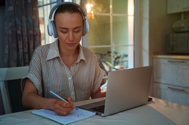 Kvinne har på hodetelefoner og skriver i en notatbok ved siden av en laptop