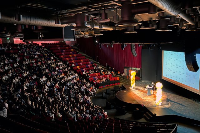 Fullsatt auditorium der en foredragsholder presenterer på en scene foran et stort publikum.