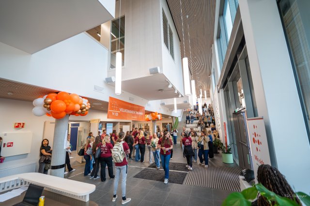 Studenter samles i en lys og moderne inngangshall under studiestart, dekorert med oransje ballonger og et velkomstbanner fra VID.