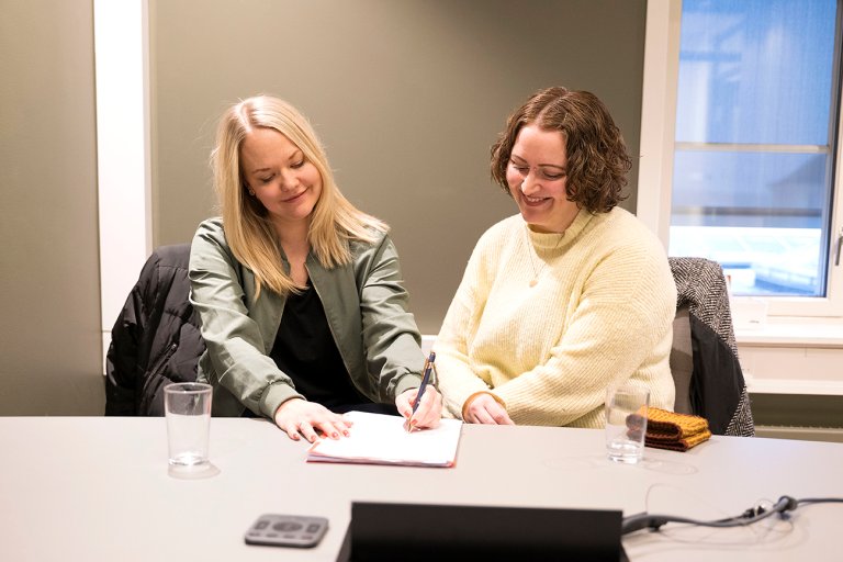 Ann Eline og Synnøve sitter på møterom og samarbeider over en notatbok. 
