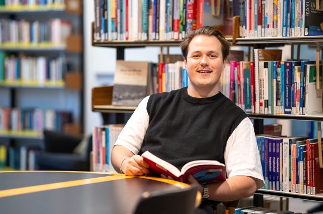 Portrett av Henrik med bok i hendene inne på bibliotek.