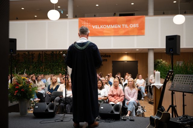 A speaker stands on a stage and welcomes new students, while an audience is gathered in front of a banner with the text "Welcome to us"