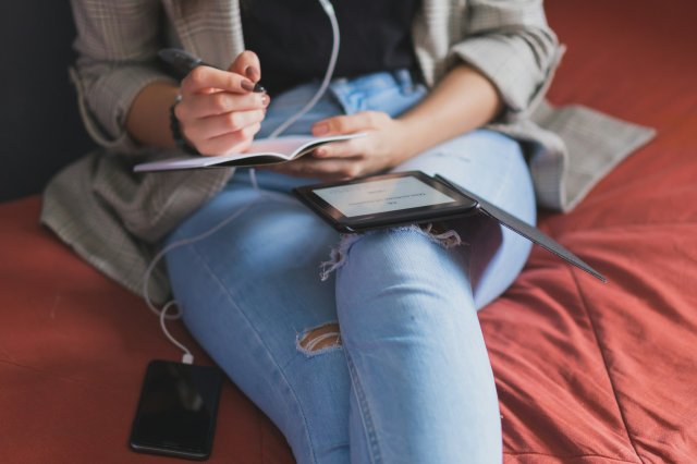 A woman sitting on a bed with an e-book reader taking notes.