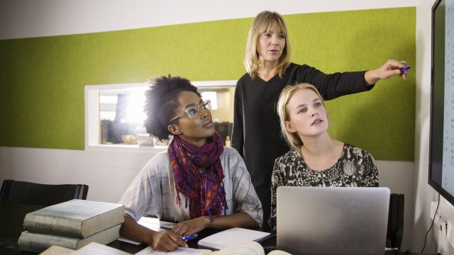 En veileder står bak og viser to voksne studenter noe på en tavle. De har bøker og datamaskin oppslått.