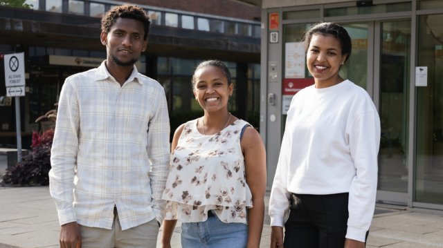 Tre studenter står på rekke og smiler til kamera