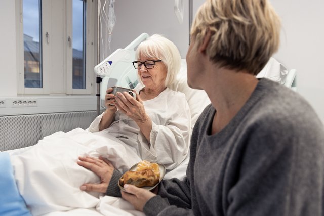 Gammel dame i sykeseng drikker noe varmt. Ved siden av sitter en person med en tallerken med mat.