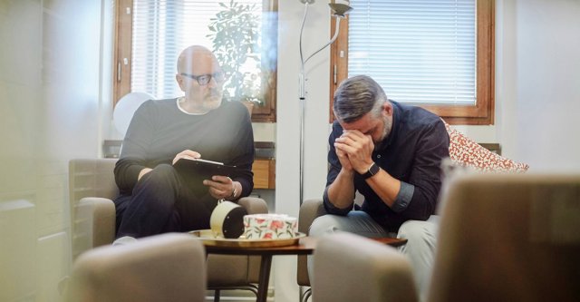To menn i et samtalerom. Den ene noterer mens den andre sitter med hodet i hendene.
