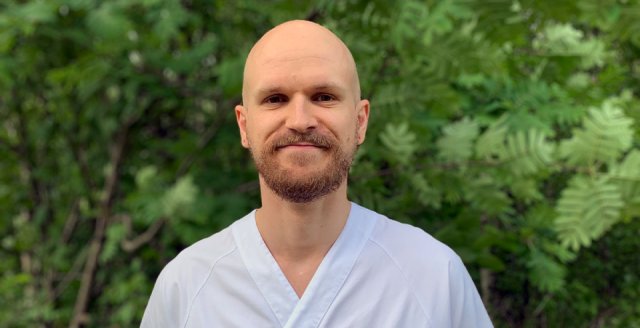 Skallet mann med skjegg og sykepleieruniform står foran en grønn busk.