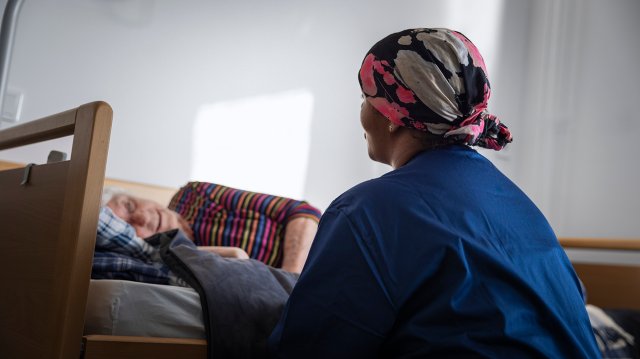 Eldre kvinne ligger i seng og helsearbeider sitter ved sengekanten og snakker med henne. 