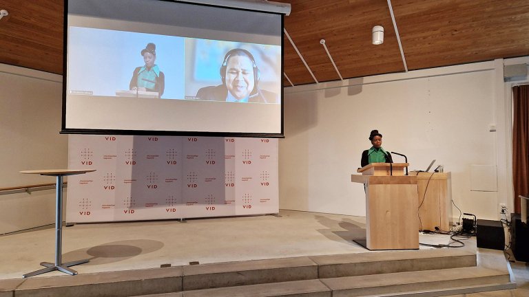 Thokozile står på talerstol foran presentasjon der man ser henne selv og Jacques.