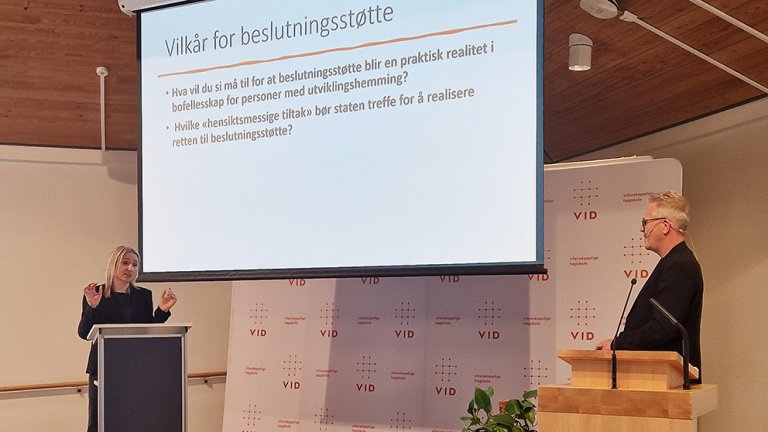 Sadeta demic på talerstolen i forelesningssal med powerpoint bak seg. 