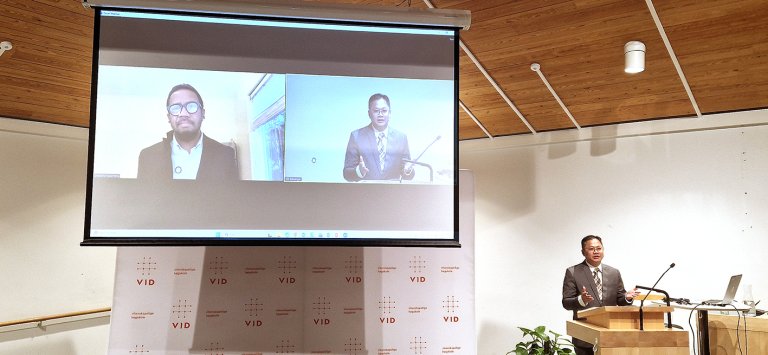 Saputra står på talerstol med førsteopponent på storskjerm bak. 