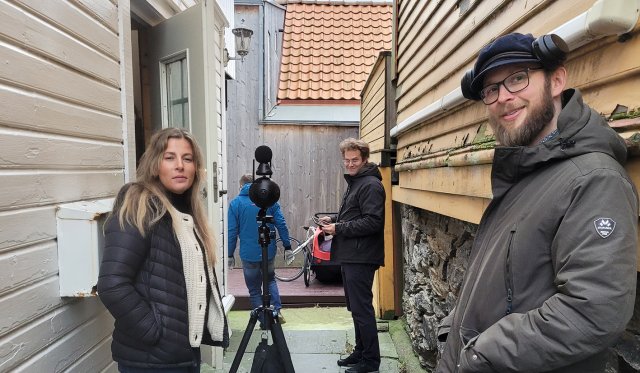 Bilde fra filminnspilling. Tre personer står utenfor et hus og ser inn i kamera.