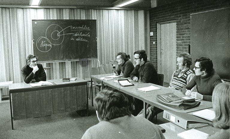 Klasserom med lærer ved kateteret og fire studenter sittende langs en vegg bak pulter. 