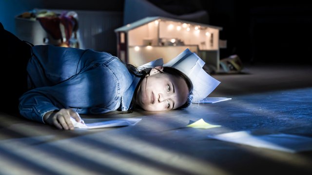 Bilde av kvinne liggende på gulvet omgitt av papirer og med et trist ansiktsuttrykk. 