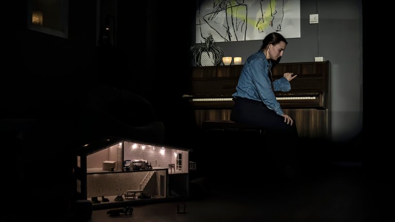 kvinne sitter i stue ved piano og ser trist i gulvet. 