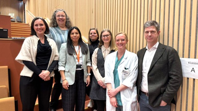 bilde av sju personer stående i forelesningssal. De ser i kamerat og smiler. 