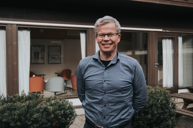 Mann med grått hår og briller poserer foran et hus