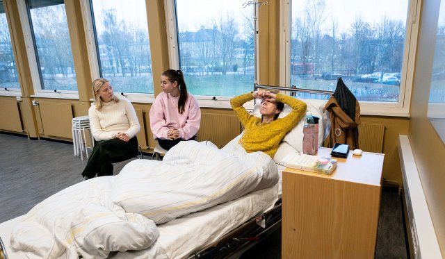 En deprimert kvinne ligger i sykehusseng. To studenter sitter ved siden av og snakker med henne.