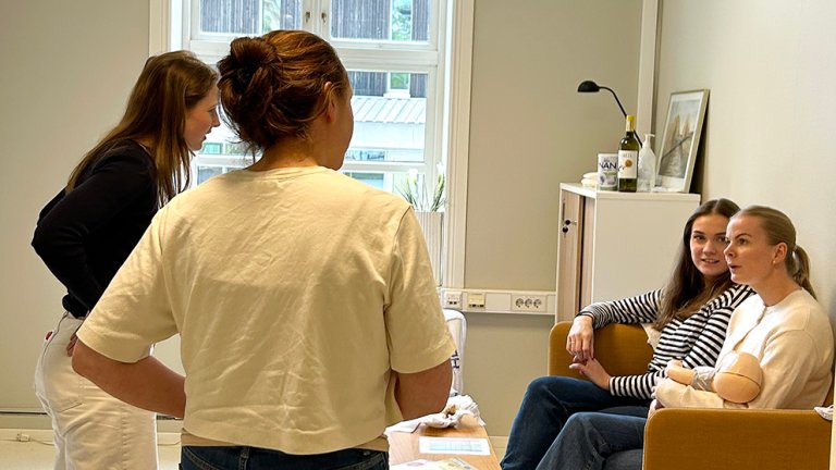 To stående studenter snakker med to studenter sittende i sofa. Den ene har en baby i armene. 