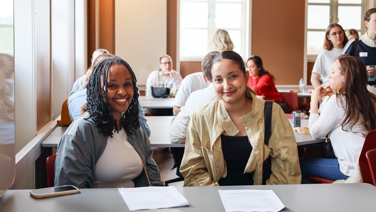 To studenter sitter ved pult i klasserom. Ser i kameraet og smiler.