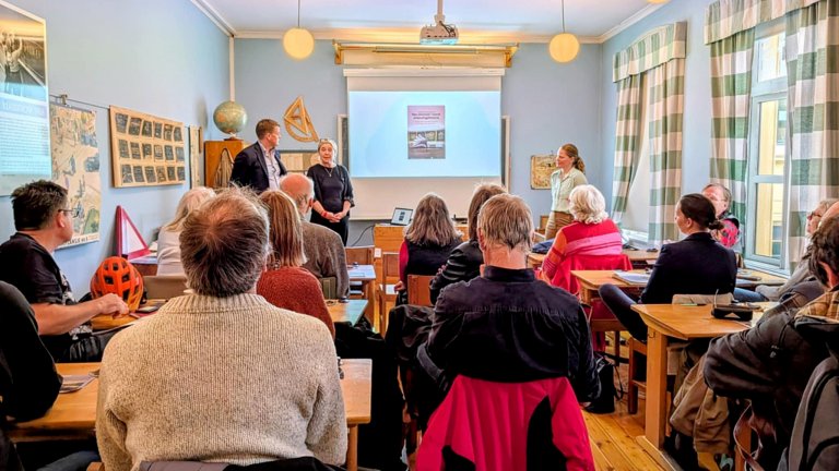 Bilde av de to forrest i klasserom med publikum på pulter. 