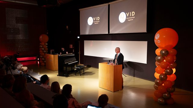 Rektor står på talerstol med ballonger ved siden av seg og VIDs jubileumslogo på skjermene bak seg.