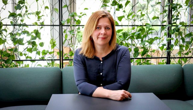 Dame med langt blondt hår sitter i en sofa og ser i kamera. Bak henne er det en klatreplanter.