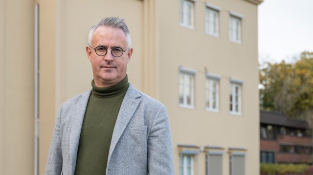 Bilde av mann i grå blazer og grønn genser foran gul bygning.