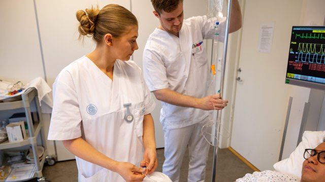 to sykepleierstudenter i uniform ved siden av sykehusseng med medstudent. 