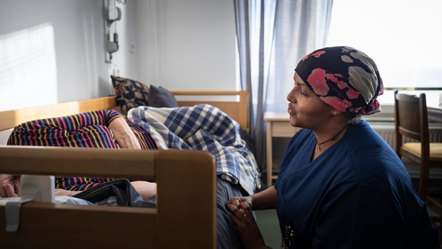 mørkhudet kvinne med hodeplagg sitter ved sengen til eldre person. 