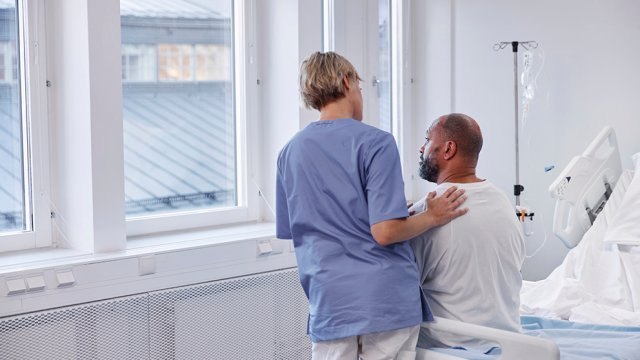 Mørkhudet pasient sitter i sykehusseng. Kvinnelig helsepersonell står ved siden av. 