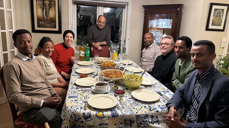En gruppe mennesker sitter rundt et bord i et hjem klare for å spise middag. Abdallah står ved enden av bordet. 