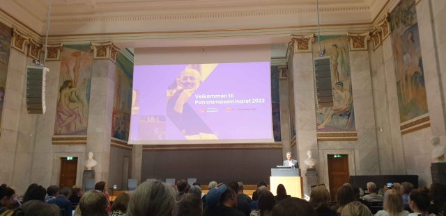 Foto av en powerpoint på lerret i et rom fullt av mennesker som ser mot lerretet.