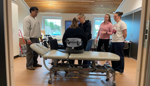 En person sitter i seng iført en bæresele. Fire mennesker står rundt.