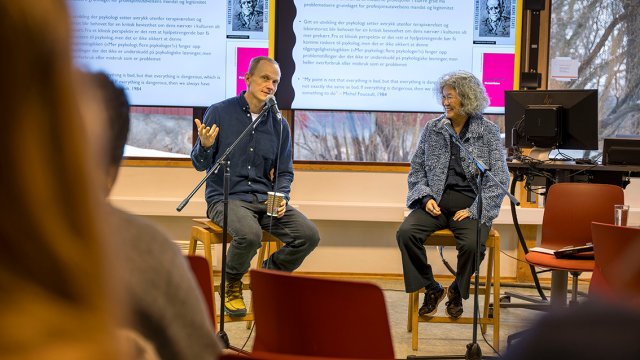 Bilde av de to sittende foran hver sin mikrofon foran publikum i klasserom.