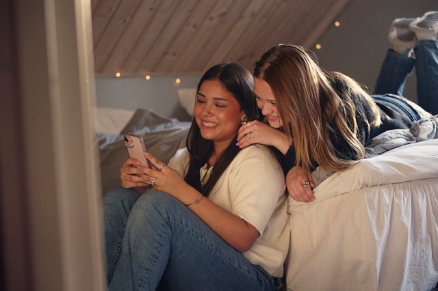 To jenter sitter og ligger på seng og smiler og ser på en mobiltelefon sammen.