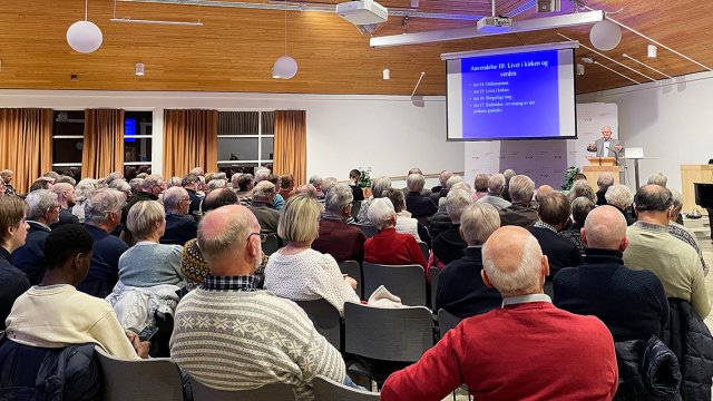 bilde tatt bakfra av sal full av mennesker med en taler foran