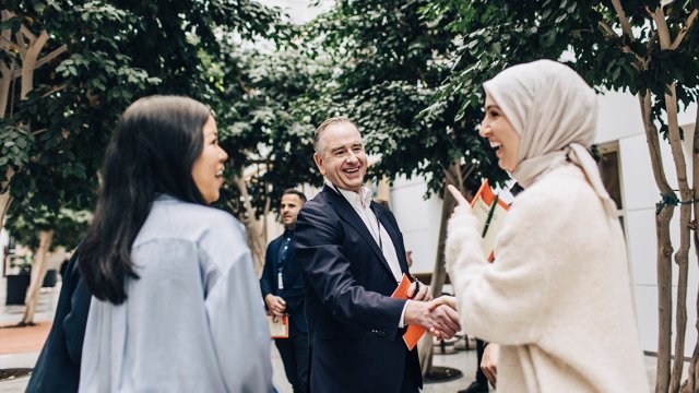 kvinne med hijab smiler og håndhilser på mann utendørs. Begge har penn og notatblokk i hendene.