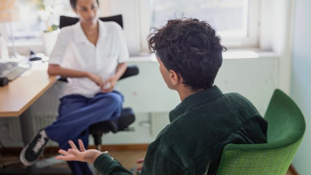 Bilde tatt bakfra av ung gutt som sitter på et kontor og snakker med en kvinne.