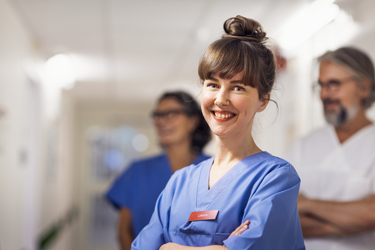 En smilende sykepleier ser i kameraet. Bak henne står en mann og en dame