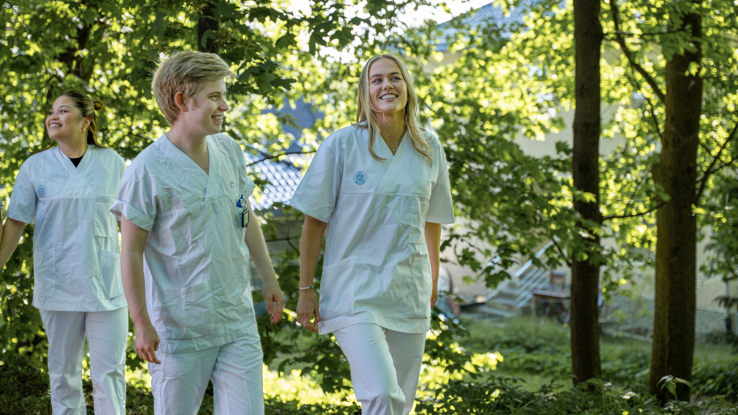 Tre sykepleierstudenter går utendørs. Rundt dem er det grønne løvtrær.