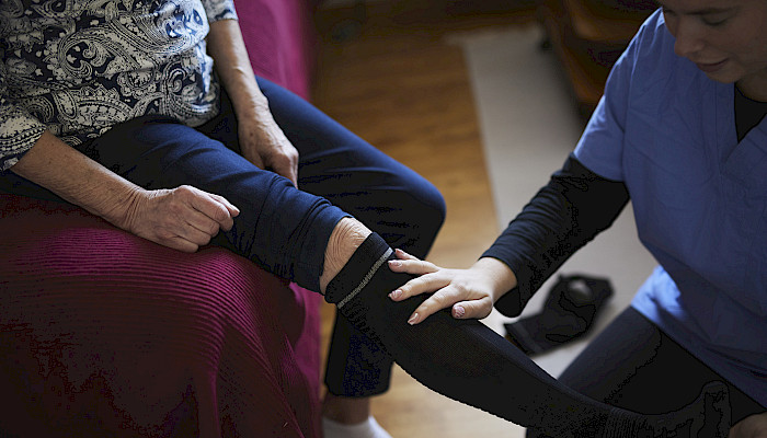 En eldre kvinne får hjelp med støttestrømpene av helsepersonell.