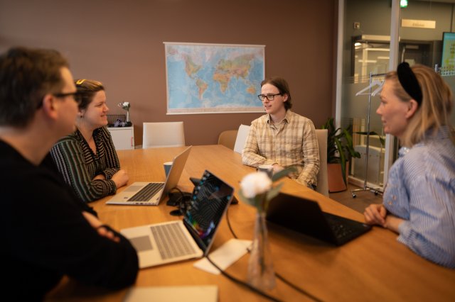 Fire studenter sitter rundt et bord og diskuterer. I bakgrunnen er det er verdenskart på veggen.