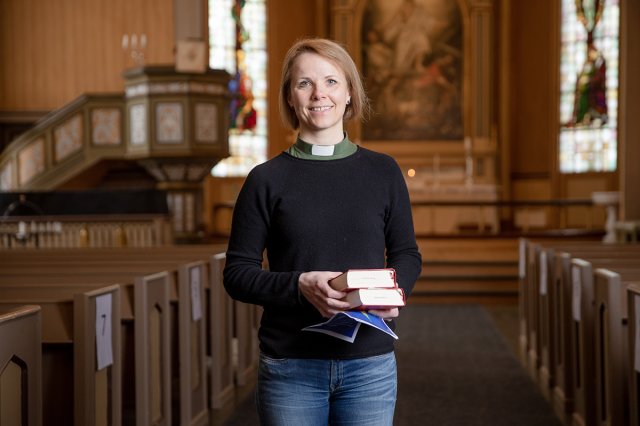 En kvinnelig prest holder bøker mens hun står i midtgangen i en kirke