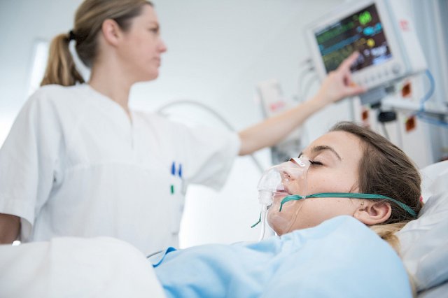 Dame trykker på en skjerm ved siden av en sykeseng. I sengen er en pasient med pustemaske.
