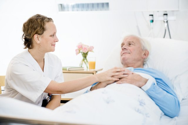 En dame sitter ved en sykeseng og holder en gammel dame i hånden