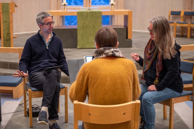 Tre voksne mennesker sitter på stoler i et kirkerom